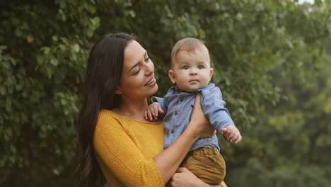 portrait shot of a smiling young woman with long dark hair holding her baby boy and looking at the camera in the park