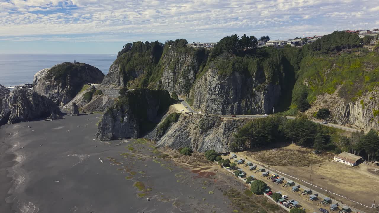 Strada sulla scogliera e area di parcheggio sopra una spiaggia di sabbia nera con onde e faraglioni al largo, Constitución, Cile, 4K