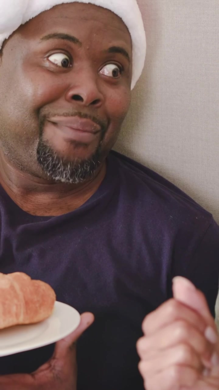 Vertical video: She leaning chatting on couch, sipping mug as man in red hat holding croissant