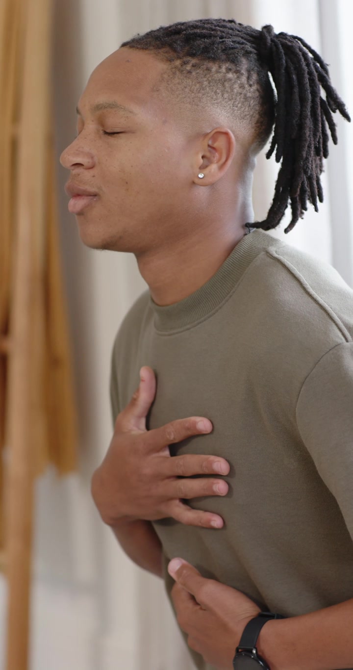 Vertical video: African man placing hands on chest and belly, breathing near window wearing watch