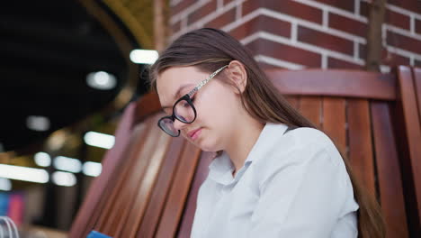 young woman wearing glasses sits on wooden bench in modern mall, deeply engrossed in book, bokeh lights in background create warm atmosphere, emphasizing focus and intellectual curiosity