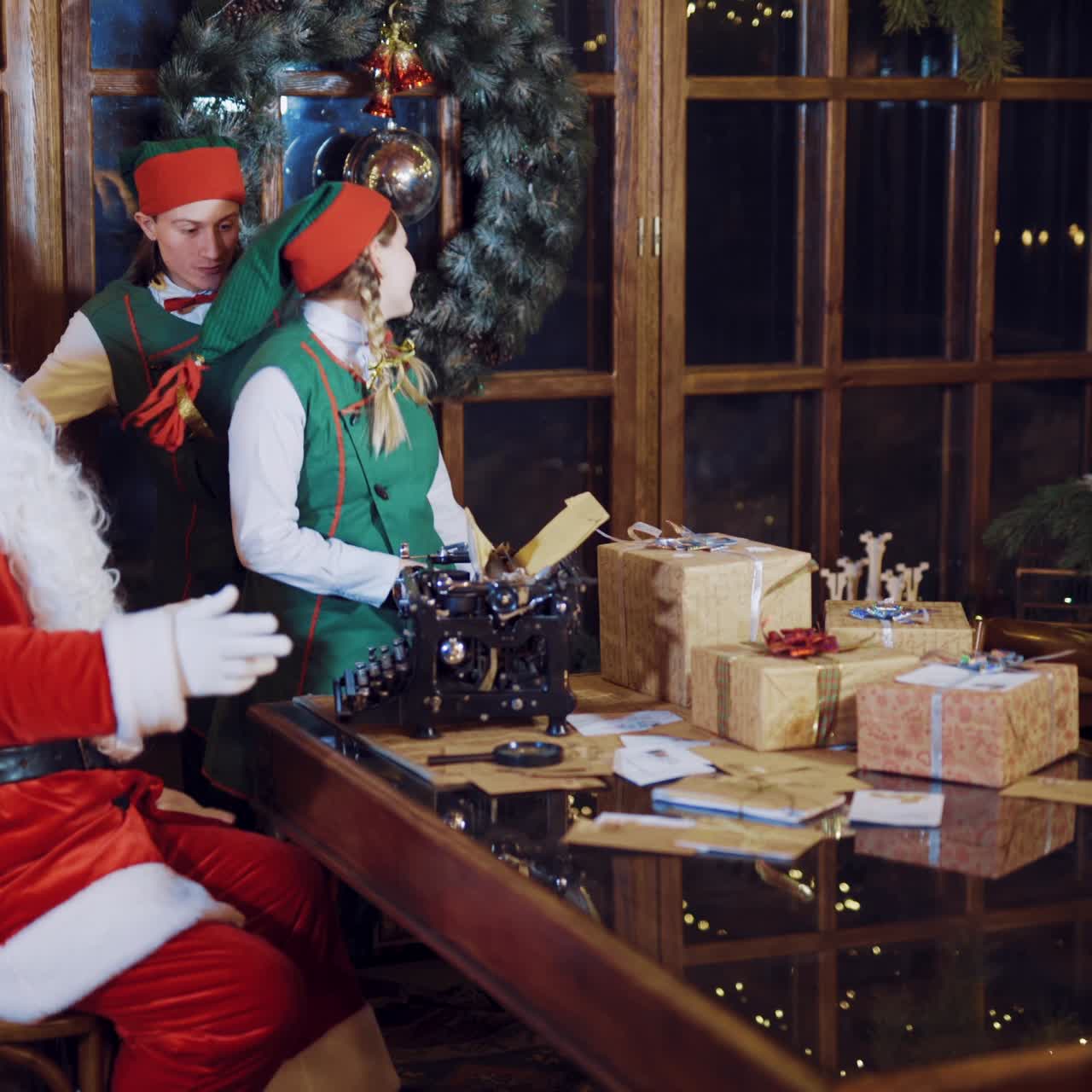 Elves in festive costumes are dancing around the table with gifts, candles and a typewriter in the presence of Santa Claus, who watching and setting the rhythm on the background of the New Year tree near the window in the room.