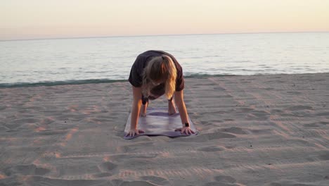 Frau,-Die-Sich-Zum-Sonnenuntergang-An-Einem-Strand-Ausdehnt-Und-Yoga-Bewegungen-Auf-Ihrer-Yogamatte-Durchführt