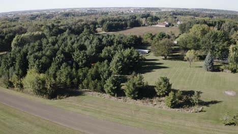 a wide rural landscape featuring rows of mature green trees, an expansive mowed grass field, and scattered houses visible in the distance under a clear sky.