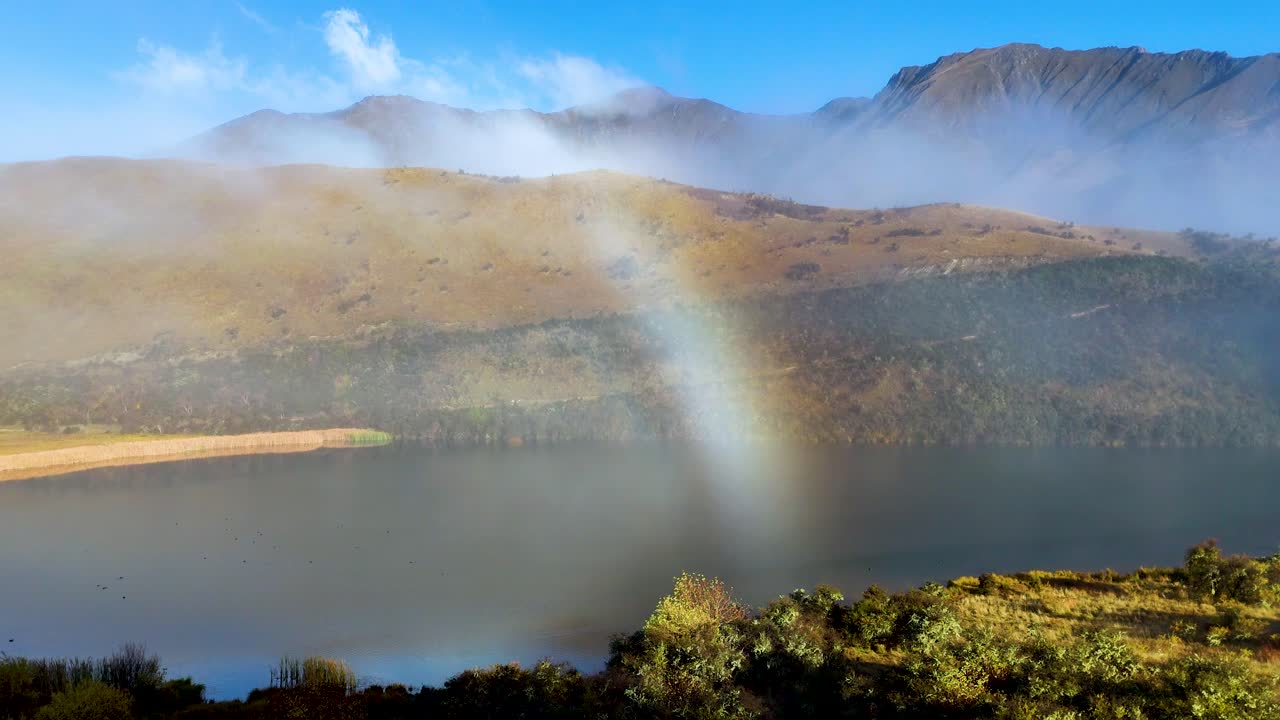 A vibrant rainbow arches over Moke Lake, surrounded by misty mountains and lush greenery, captured in bright daylight