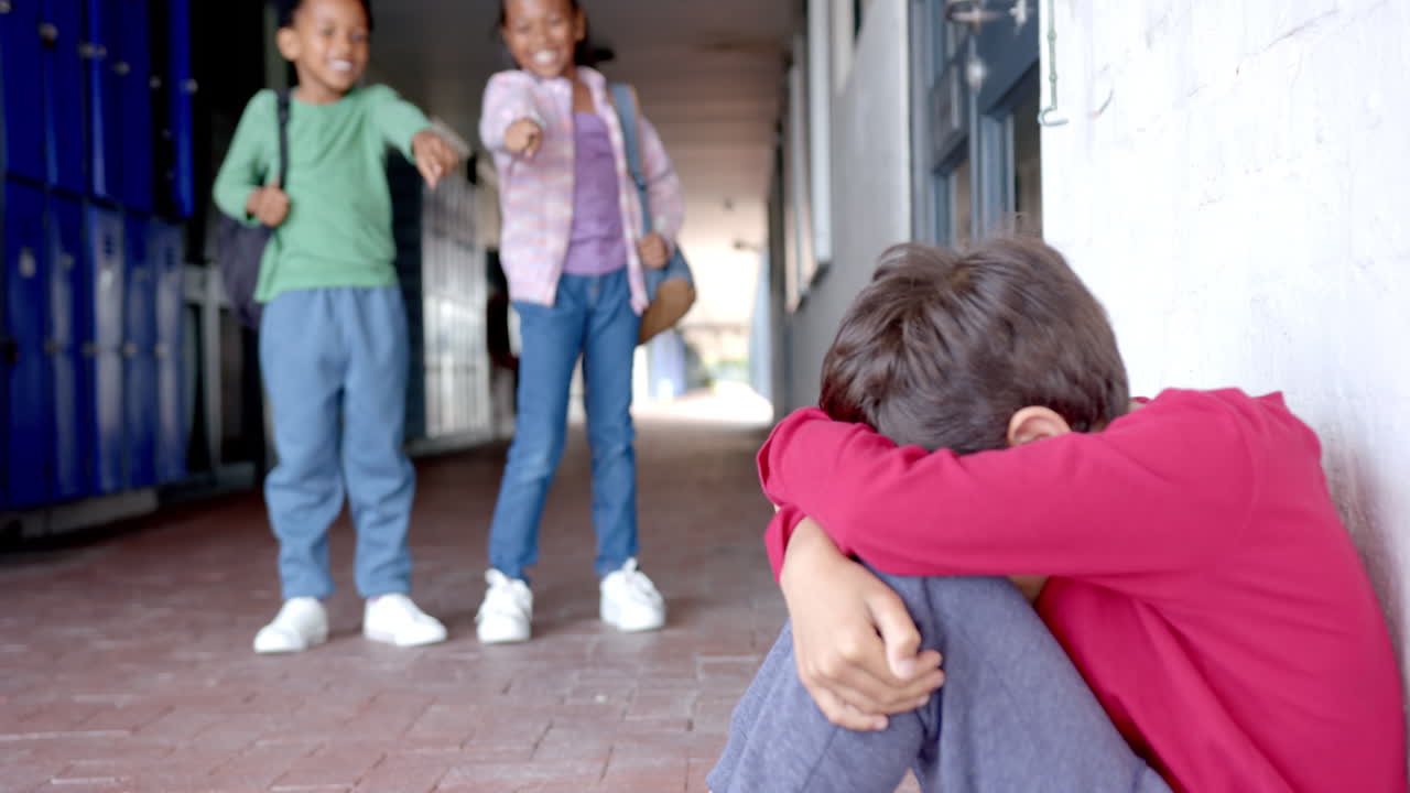 In school, two children bullying another child sitting on floor in hallway