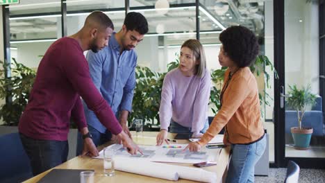 Diverse-office-colleagues-wearing-face-masks-discussing-over-blueprint-plan-at-modern-office