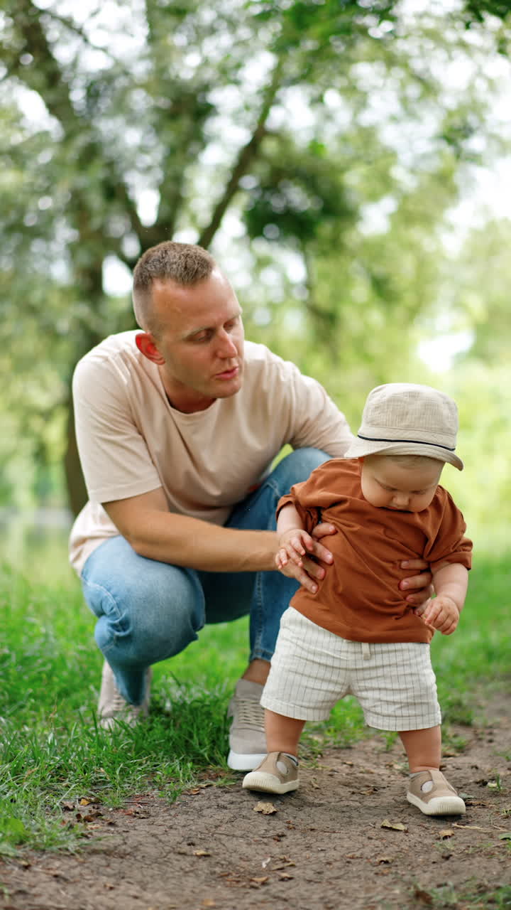 Smiling Caucasian father holds his son standing on the ground road. Dad talks to his baby boy trying to walk. Vertical video.