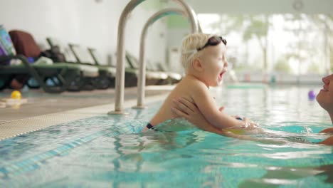 madre joven está enseñando a su bebé rubio lindo a nadar en la piscina. ella está sentada junto al agua y luego le ayuda a