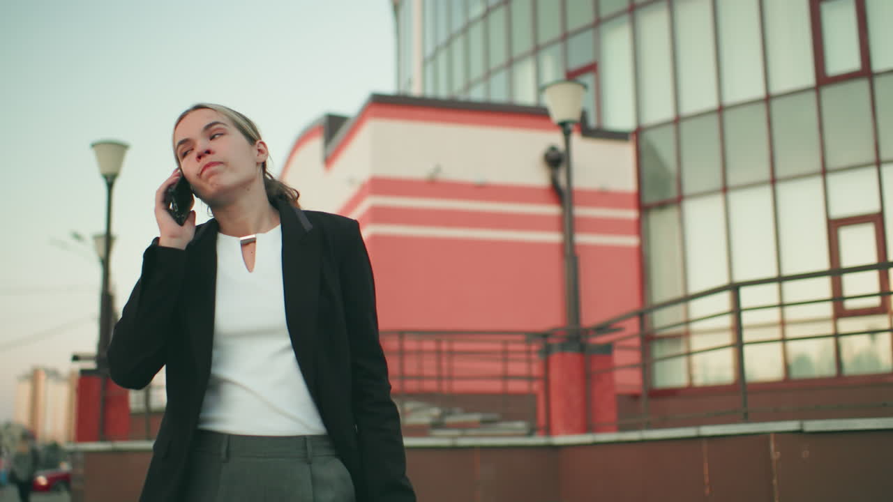 Frustrated white lady in professional outfit talking on phone looking tired and upset, walking near modern building with blurred crowd in distant background