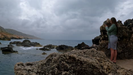 fotografía de una turista tomando fotos de las olas que se estrellan a lo largo de la playa rocosa de malta en un día nublado