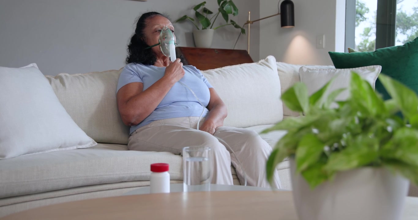 Donna anziana che inizia un trattamento respiratorio tenendo un nebulizzatore al viso a casa, con un flacone di pillole nelle vicinanze