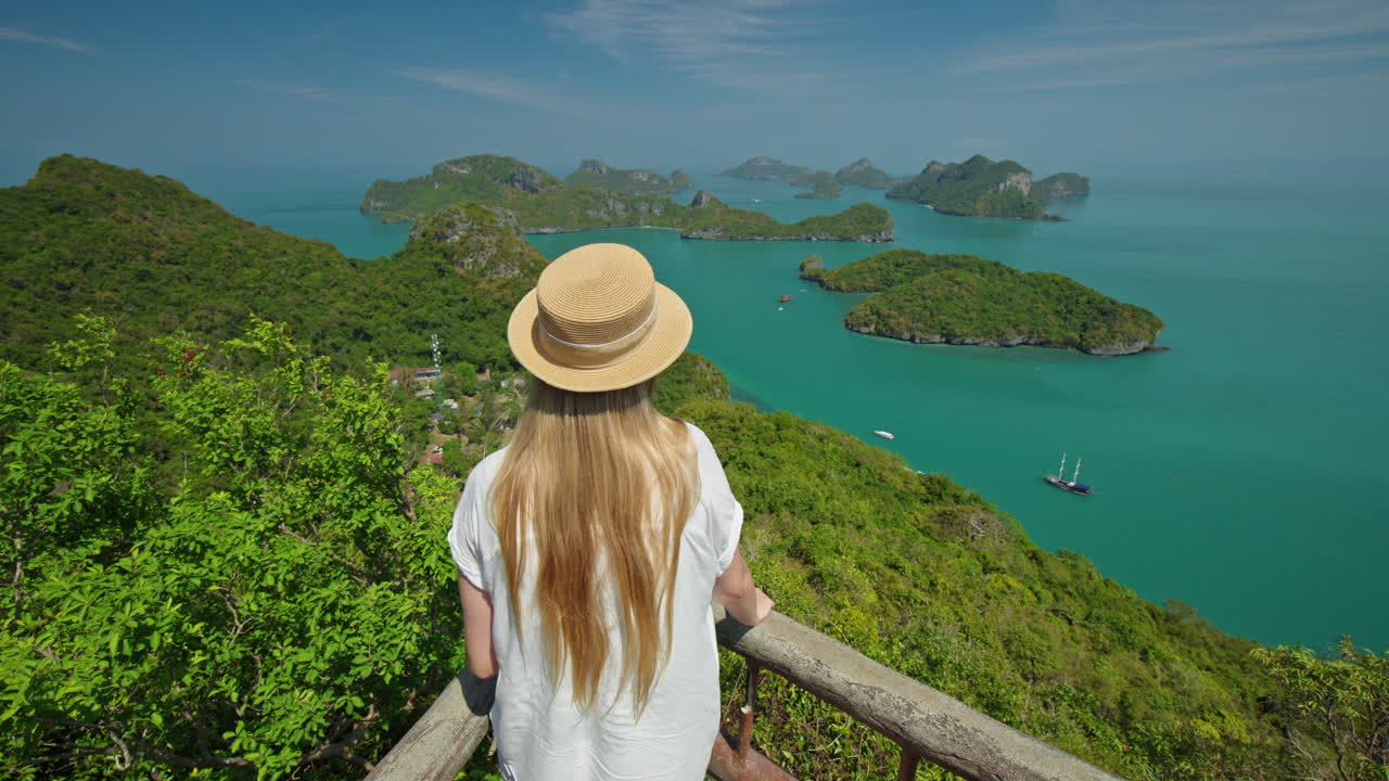 Wanita muda mengenakan topi jerami berdiri di titik pandang panorama, memandang ke laut pirus dan pulau-pulau kapur hijau di Taman Laut Ang Thong, menikmati alam tropis dan pemandangan pulau.