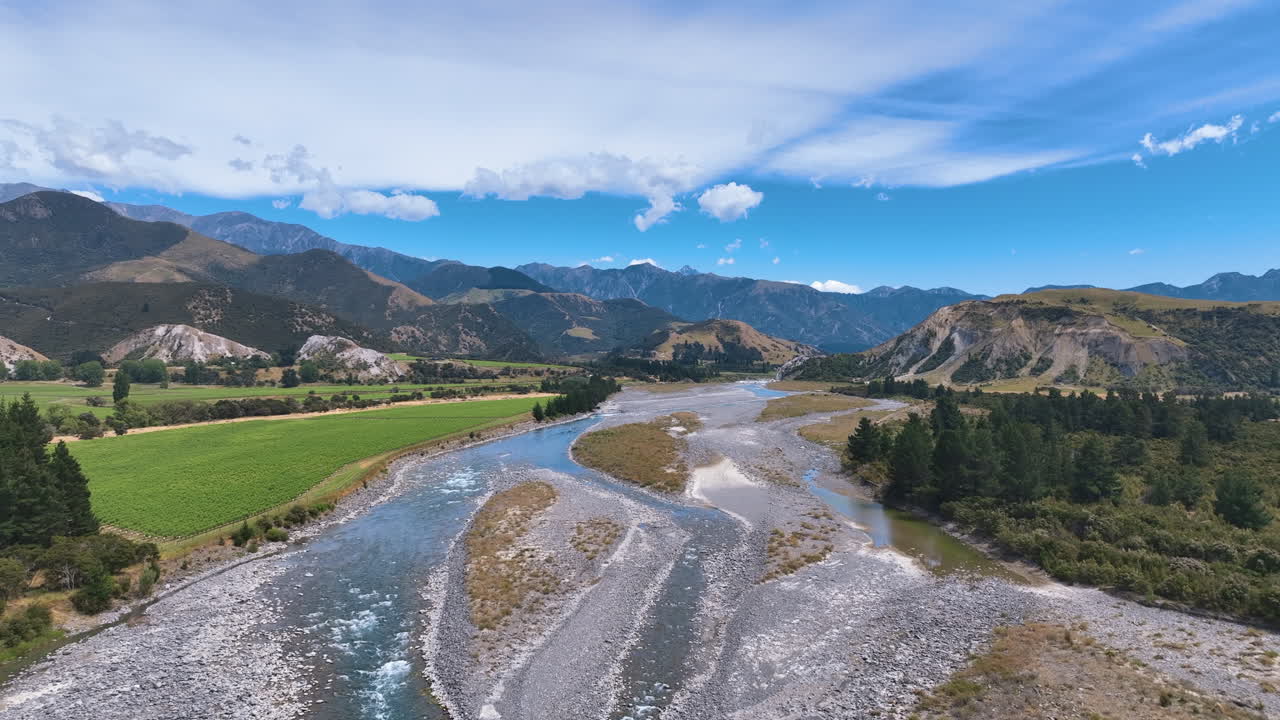 Aerial follows the cool grey blue waters of the Buller River with braided channels, flanked by green fields and conifers, snaking past hills, and flanked by huge mountains West Coast New Zealand