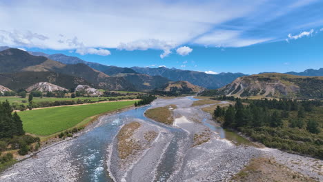 el avión sigue las frías aguas grises y azules del río buller con canales trenzados, flanqueados por campos verdes y coníferas, serpenteando más allá de las colinas, y flanqueado por enormes montañas en la costa oeste de nueva zelanda.