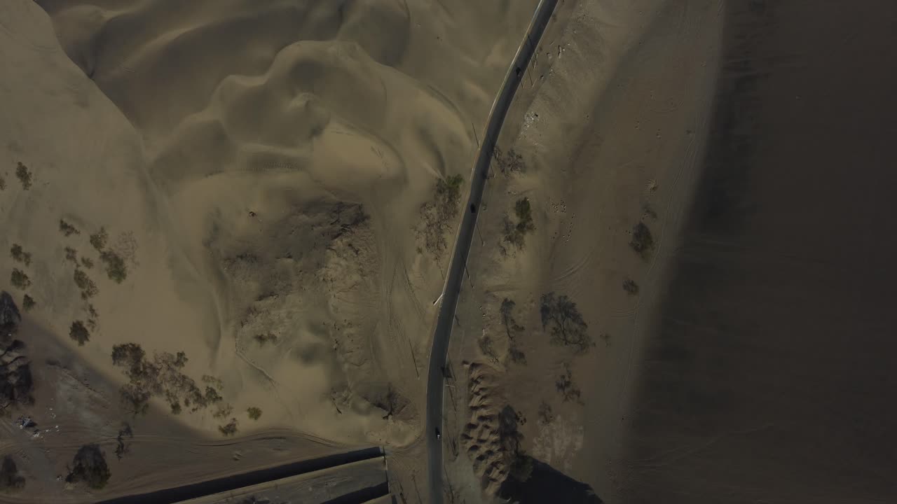 Aerial drone shot of a narrow road leading to a hidden village oasis in the middle of the desert surrounded by sand dunes. Drone hovers and tilts camera up. Located in "Huacachina" in Ica, Peru