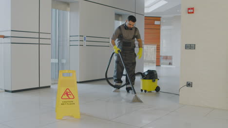 Hombre-De-Limpieza-árabe-Limpiando-Con-Máquina-De-Agua-A-Presión-Dentro-De-Un-Edificio-De-Oficinas-Detrás-De-Una-Señal-De-Advertencia-De-Suelo-Húmedo