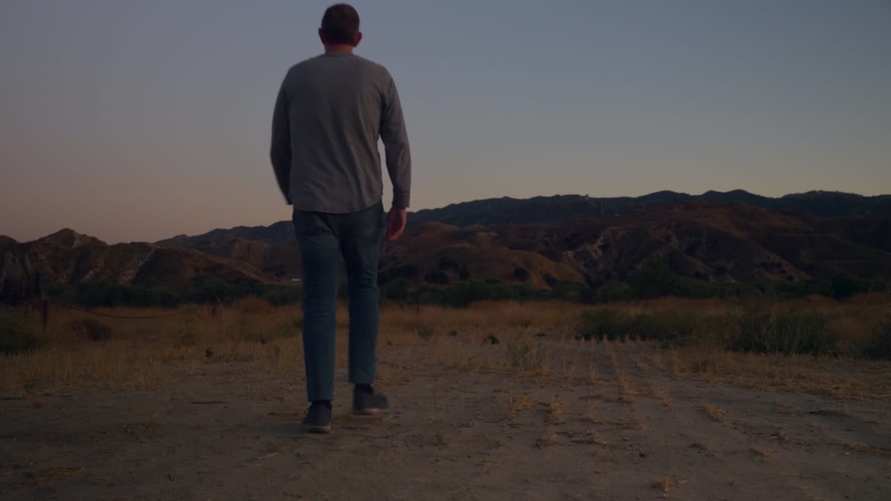 Inquadratura fissa di un uomo che entra nell'inquadratura, nella campagna californiana, al crepuscolo, sera soleggiata con montagne in lontananza