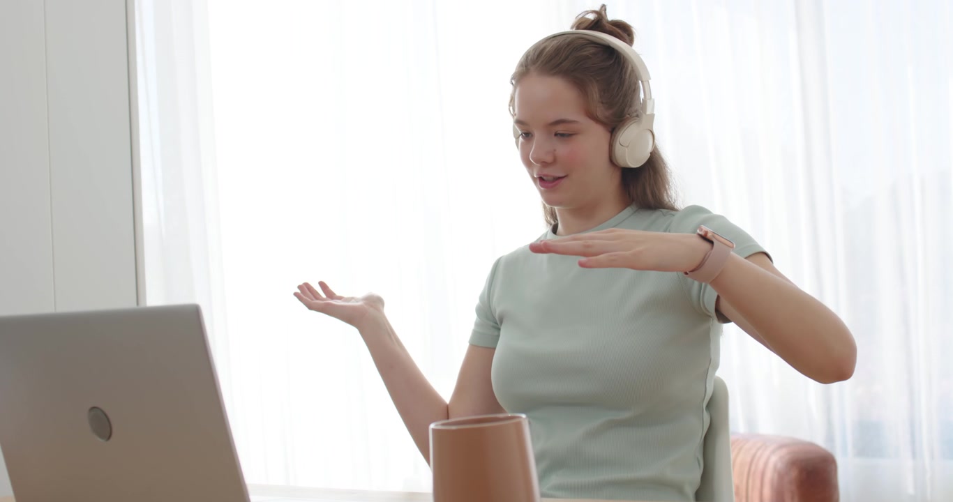Woman wearing headphones gesturing, talking in online call at home desk with laptop and cup