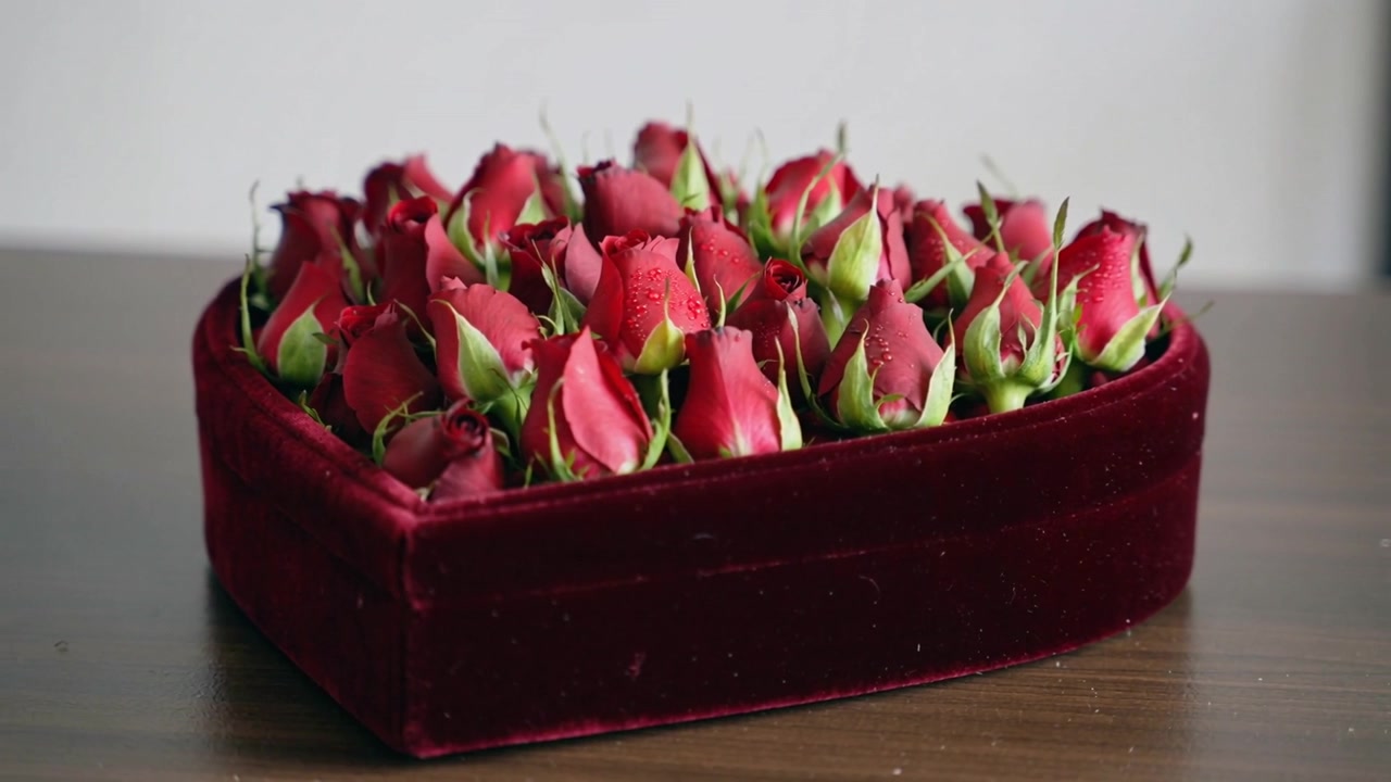 L'obiettivo si avvicina a una scatola a forma di cuore di velluto con boccioli di rosa rossi su un tavolo di legno che mostra goccioline