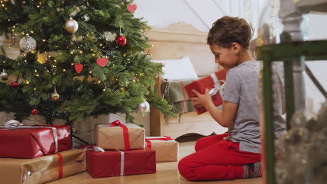 excited boy shaking and opening presents by tree on christmas morning