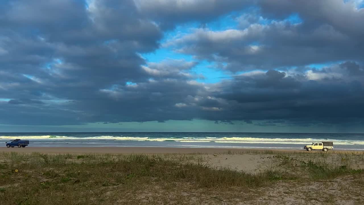 los vehículos todoterreno 4x4 conducen por una playa con una tormenta dramática que oscurece el horizonte, en un contraste de condiciones climáticas