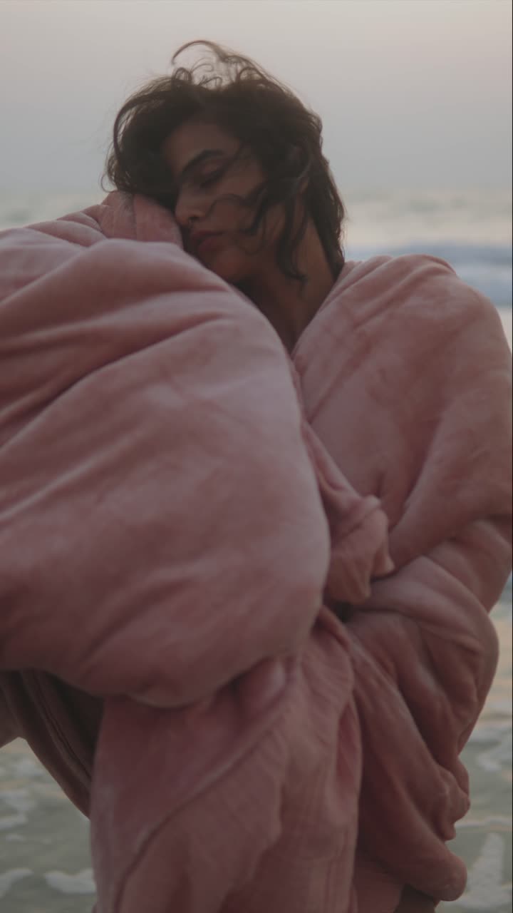 Persona rilassata avvolta in una coperta rosa in riva al mare all'alba, con un'aria serena