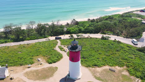 Red-and-White-Light-House-Aerial-Drone-Reveal-of-Ocean-and-Beach-in-Cape-Cod-Massachusetts-with-People-on-Beach-and-Parking-Lot