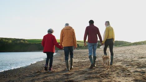 Ein-älteres-Paar-Hält-Sich-An-Den-Händen,-Während-Es-Mit-Seinem-Erwachsenen-Nachwuchs-Im-Winterlichen-Strandurlaub-An-Der-Küste-Entlang-Spaziert