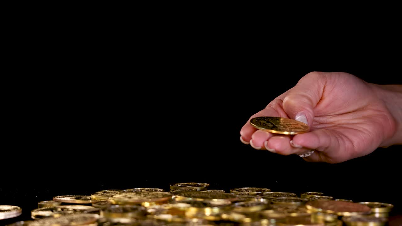 Human hand tosses and flips gold coins above pile, dramatic lighting, black background, close-up