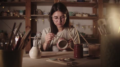 young woman potter in glasses drawing a design on the ceramic mug with a brush