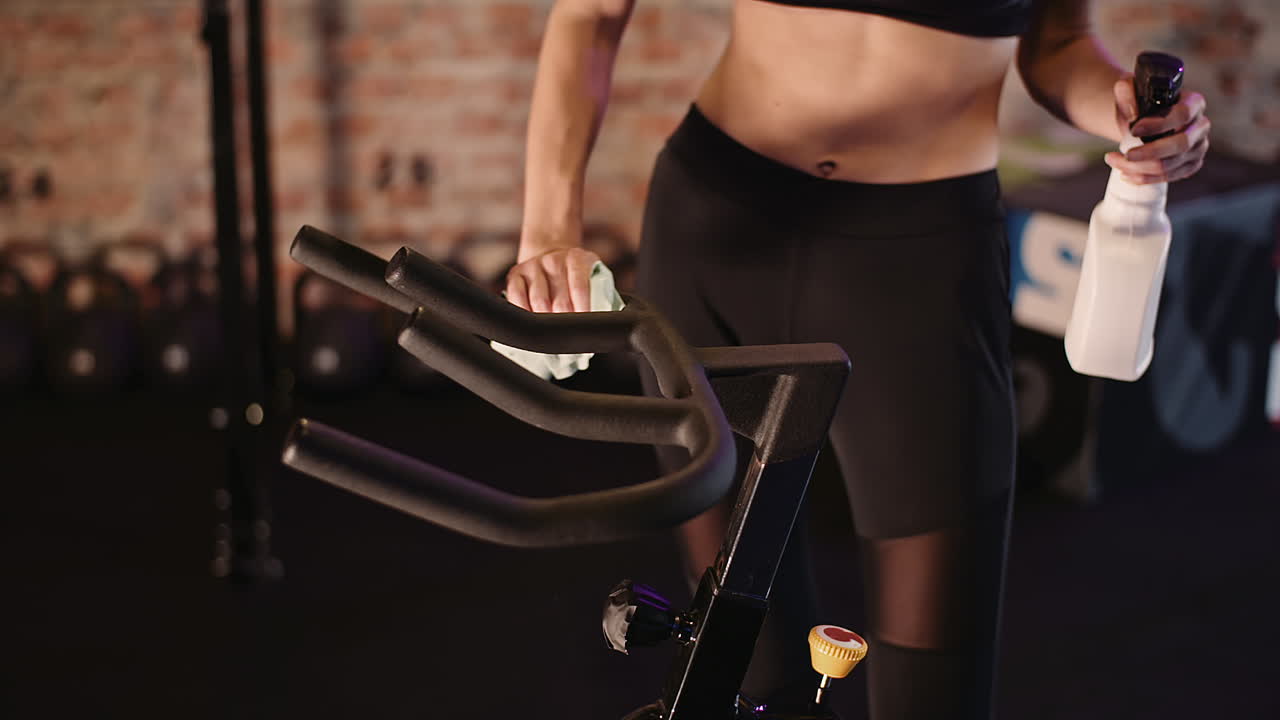 Inquadratura a mezzo busto di un'istruttrice che pulisce il manubrio di una cyclette in palestra.