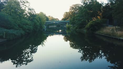 Drohnenantenne,-Die-Sich-Bei-Sonnenaufgang-Rückwärts-über-Den-Fluss-In-Einem-Park-Bewegt,-Wobei-Eine-Brücke-Auf-Dem-Wasser-Reflektiert-Wird