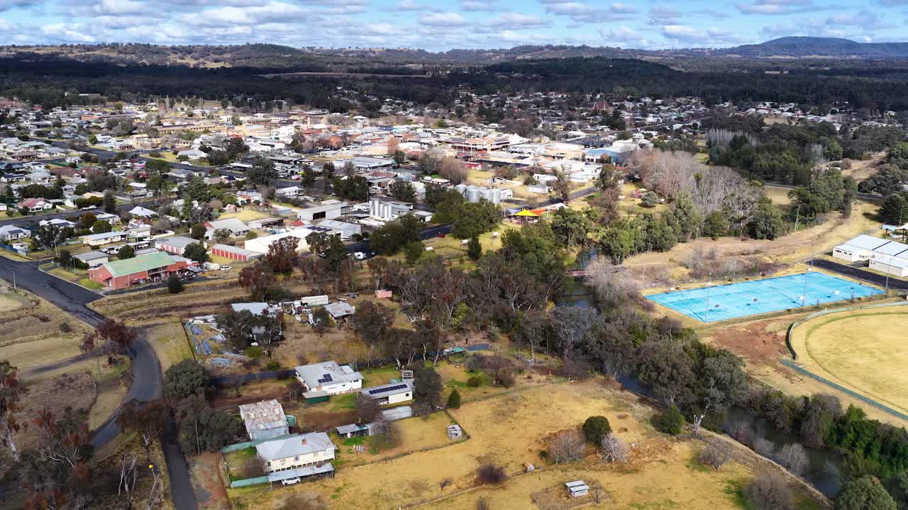 Drone footage glides above suburban homes, sports fields, and tree-lined streets in Coonabarabran, NSW, under overcast daylight with steady, forward camera movement