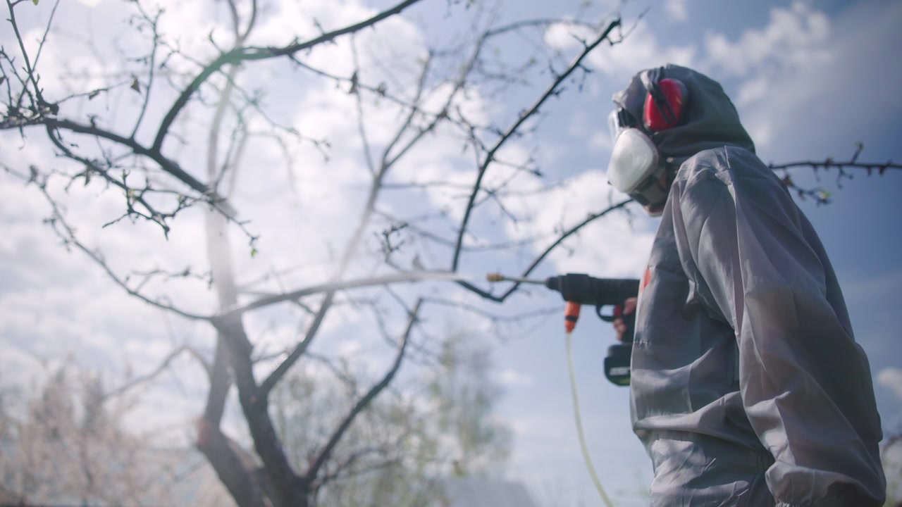 Profilo laterale di un lavoratore con tuta protettiva grigia con cappuccio, visiera trasparente, maschera respiratoria e cuffie antirumore rosse che tiene un ugello irroratore verso rami di alberi spogli in un tranquillo ambiente esterno.
