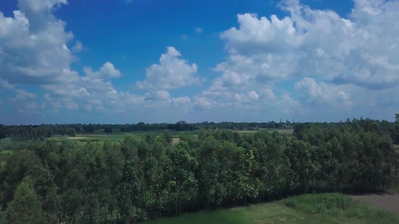 Drone advances over patchwork rural Indian fields of green paddies and brown plots toward distant lush treeline along winding river under expansive blue sky with scattered white clouds