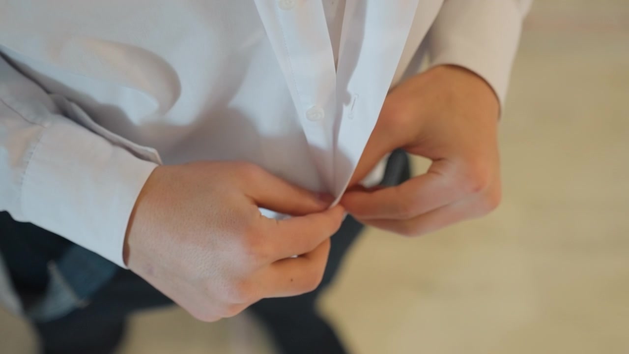 Uomo caucasico ripreso dall'alto che si abbottona la camicia, mani concentrate in una mattinata aziendale, camicia bianca elegante, pantaloni scuri, sedia da ufficio, luce ambientale brillante, nervosismo pre-colloquio, abbigliamento professionale, atmosfera dinamica.