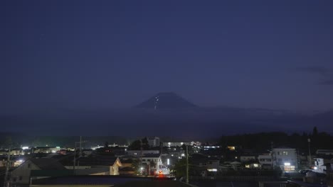 Tranquil-nighttime-scene-showcasing-the-iconic-Mount-Fuji-in-Japan,-with-a-lively-village-below,-aglow-with-the-movement-of-cars-and-vibrant-night-activities