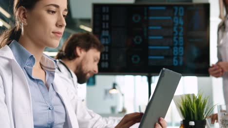 Close-up-view-of-Caucasian-female-doctor-typing-on-tablet-and-looking-at-digital-screen-presentation-sitting-at-meeting-with-multiethnic-doctors-group