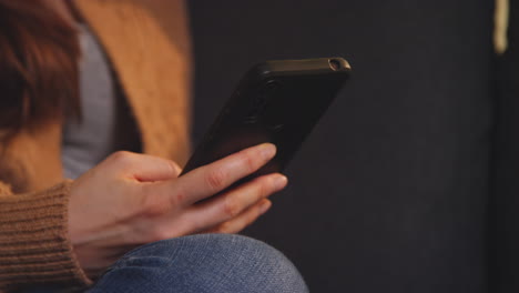 Primer-Plano-De-La-Mano-De-Una-Mujer-Sentada-En-Un-Sofá-En-Casa-Usando-Un-Teléfono-Móvil-Para-Revisar-Los-Mensajes-De-Las-Redes-Sociales-Y-Desplazarse-En-Línea