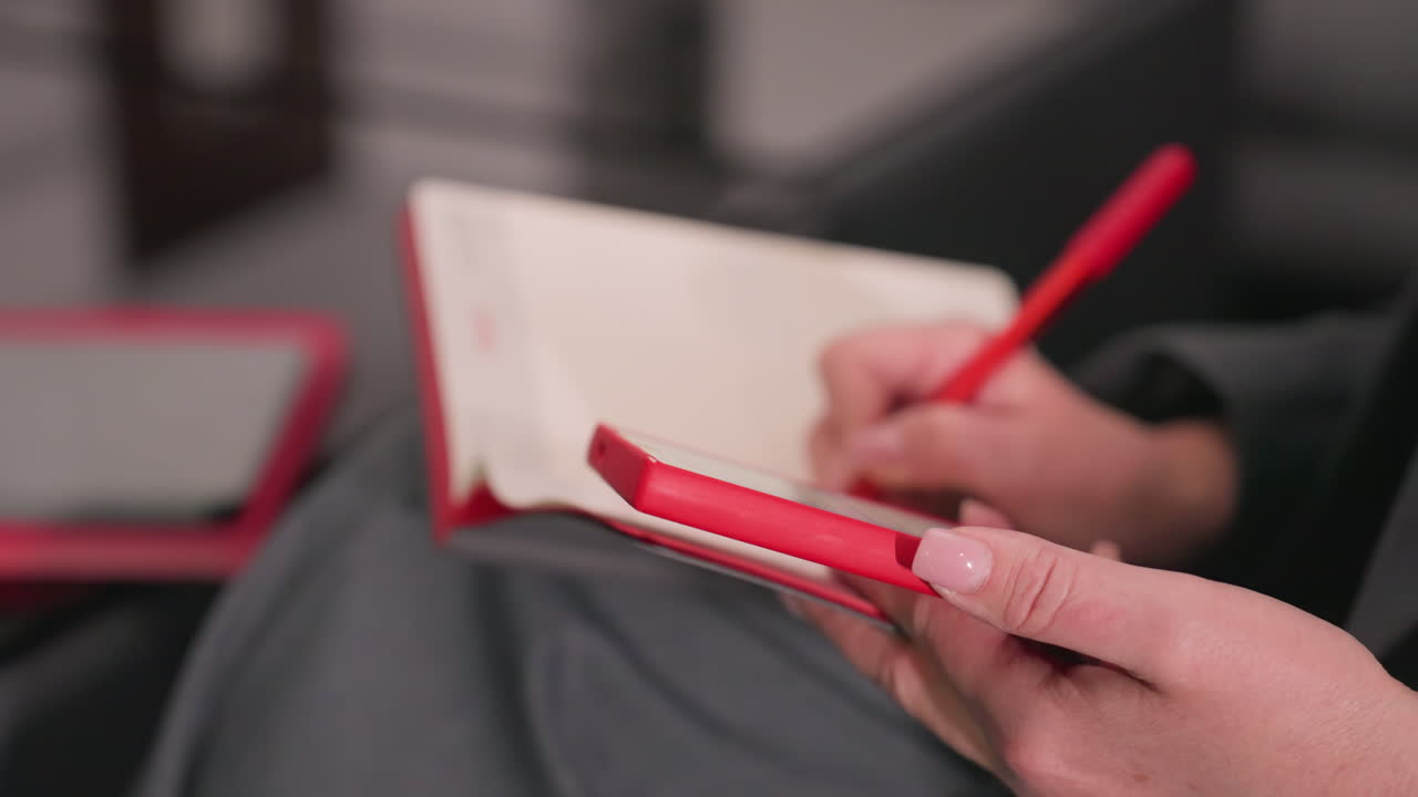 Close-up of person holding red phone in one hand and writing with red pen on notebook, showcasing focus on task at hand. Ideal for business meetings, office work, and productive sessions