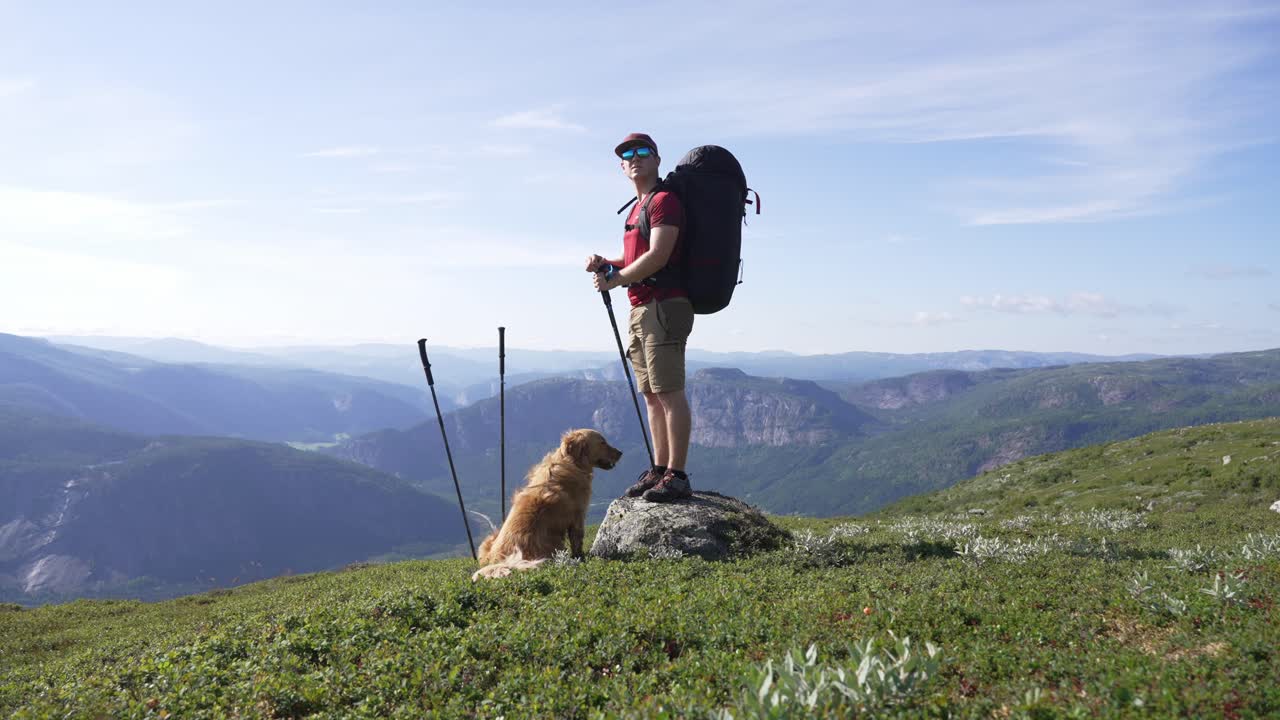 A hiker with a backpack and dog stands on a rock in scenic Norway mountains