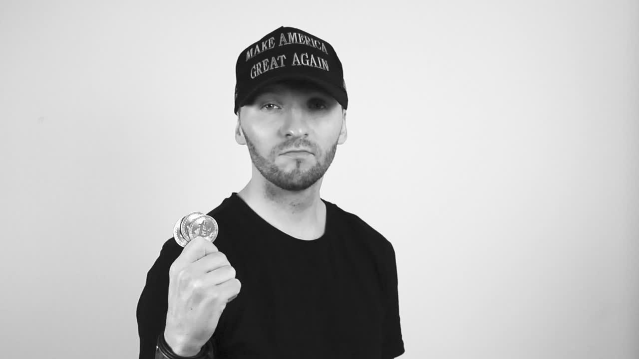 Grayscale footage of a tattooed man with short beard and a dark black MAGA or Make America Great Again hat holding up three shiny and reflective bitcoin cryptocurrency coin and watching them, happy.