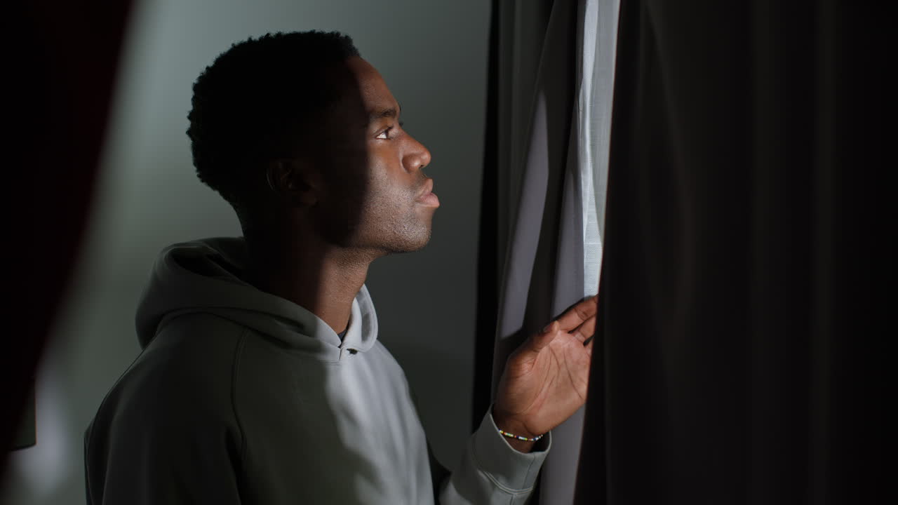Unhappy And Depressed Young Man At Home Opening Curtains And Looking Out Of Window