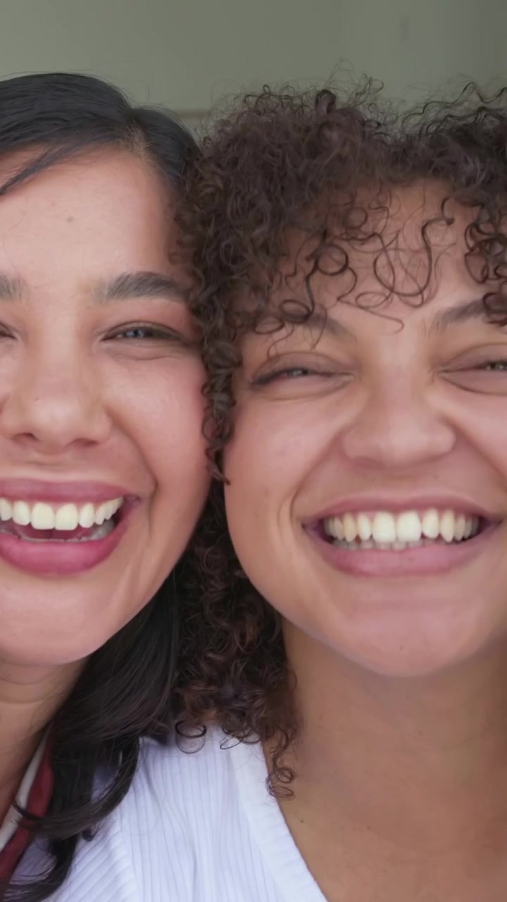 Video vertical: Alcanzando el teléfono, dos amigas con tops óxido y blancos echándose hacia atrás, tomándose una selfie en casa.