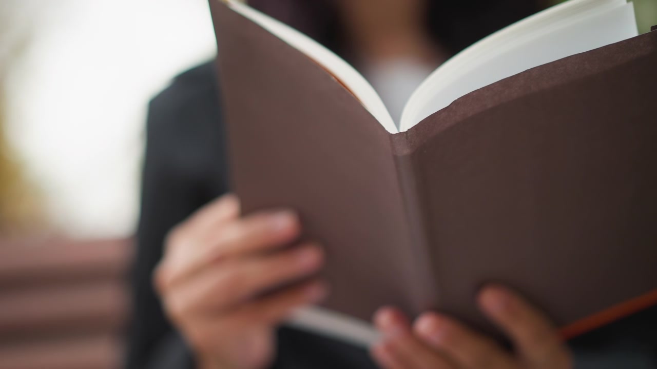 Primo piano di una donna che tiene e legge un libro aperto con pagine bianche. La donna indossa abiti scuri, concentrandosi sulle mani che stringono un taccuino rilegato in copertina rigida marrone. La luce naturale soffusa illumina la scena.