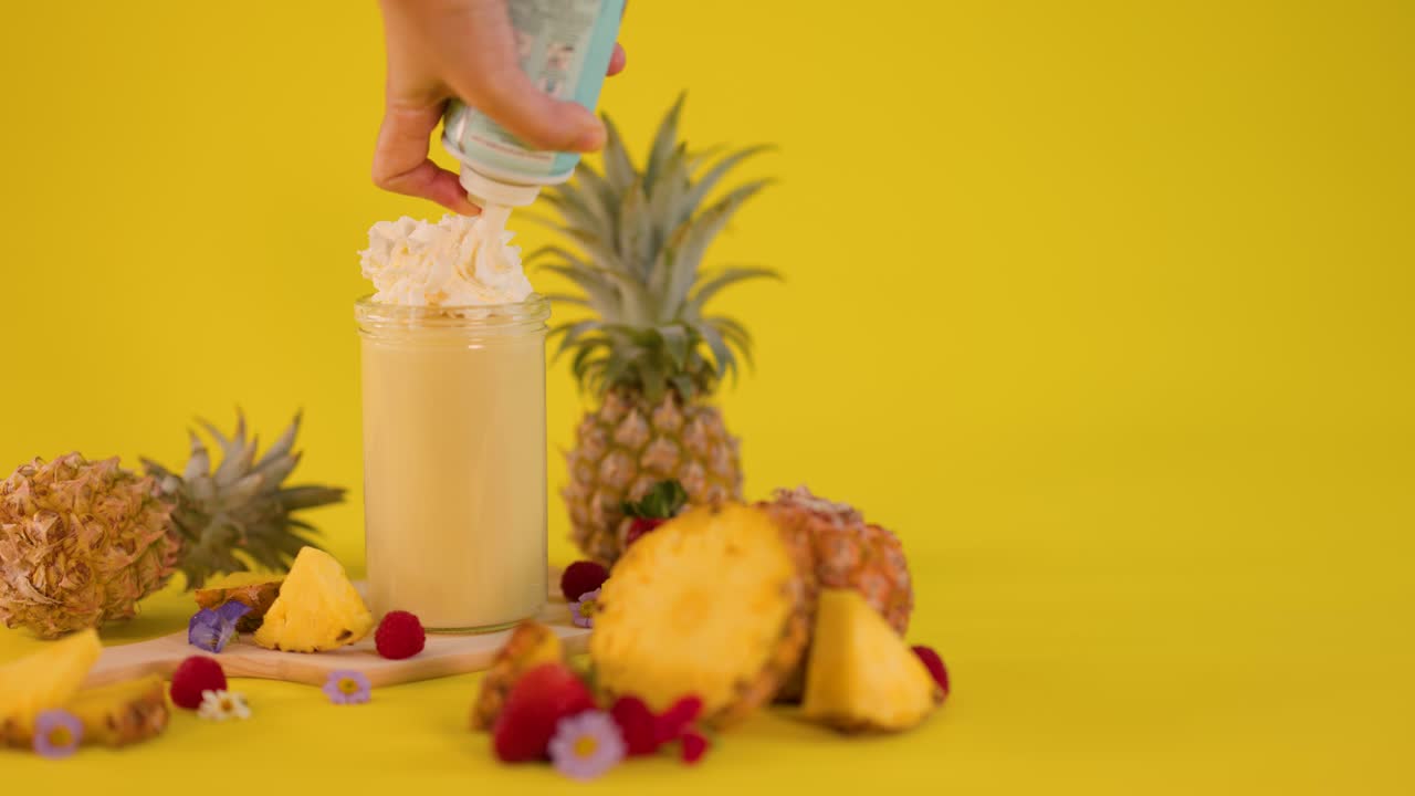 Hand adds whipped cream to pineapple smoothie surrounded by tropical fruit on vibrant yellow background