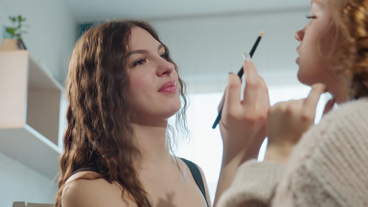 Friends Applying Makeup, Warm Apartment Setting With Friends Applying Makeup And Sharing Laughter, Caucasian Friends Enjoy Playful Grooming And Blush Application Under Cozy Apartment Lighting
