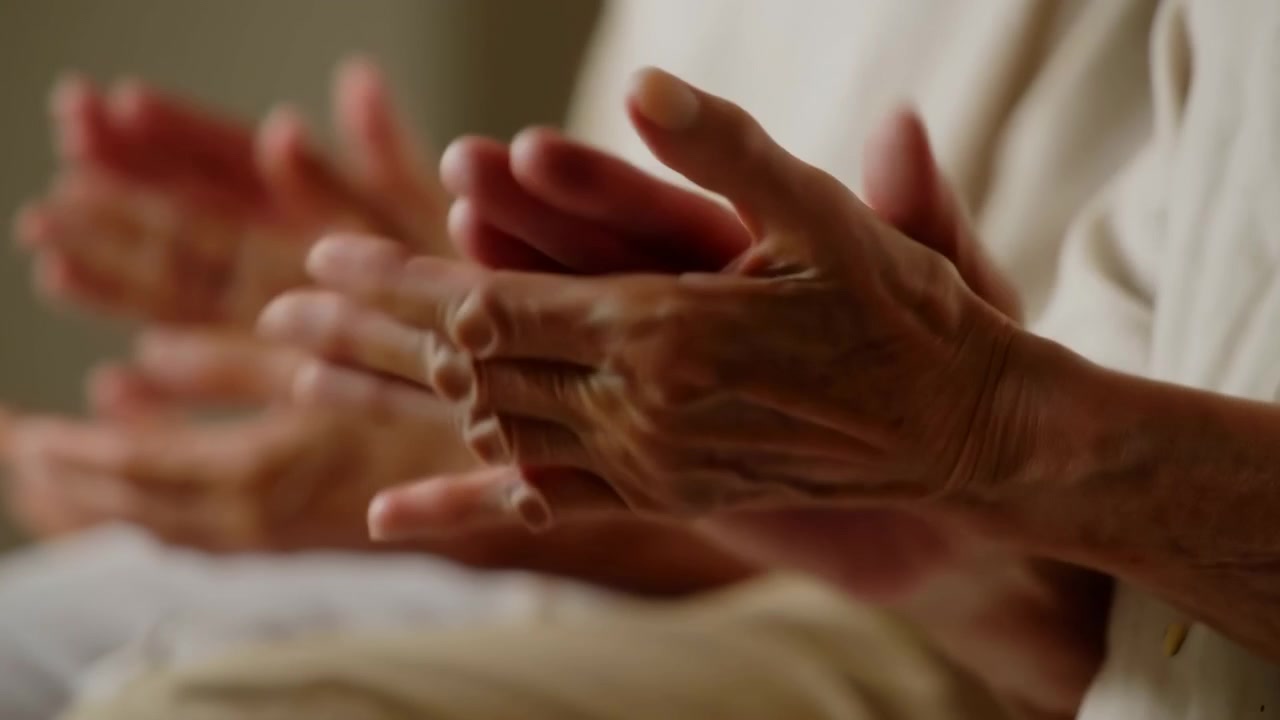 Primo piano panoramico di mani anziane che applaudono all'altezza del petto in una calda luce interna, con sfocatura a strati e movimento rilassato che creano un senso di ritmo delicato, supporto, connessione e apprezzamento condiviso.