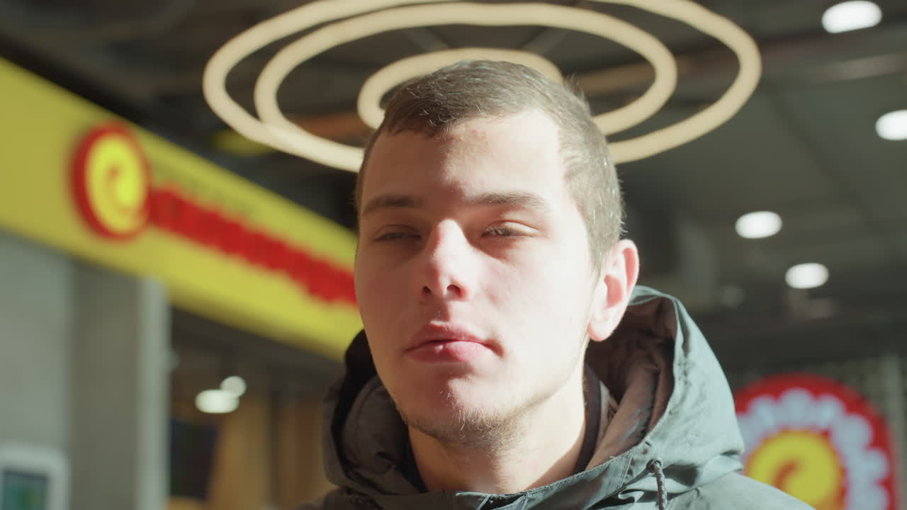 Uomo bianco sotto luci ad aureola, un ritratto urbano contemplativo con calde insegne commerciali. Illuminazione ad anello interna, cappuccio della giacca invernale, bokeh morbido e un'espressione solenne catturata in primo piano.